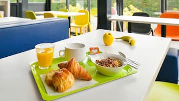 Bandeja de desayuno con cruasanes, un tazón de cereal, un vaso de jugo de naranja y un plátano sobre una mesa en una habitación luminosa con ventanas grandes y sillas amarillas.