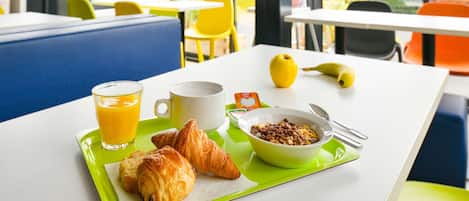 A breakfast tray with croissants, a bowl of cereal, a glass of orange juice, and a banana on a table in a bright room with large windows and yellow chairs.