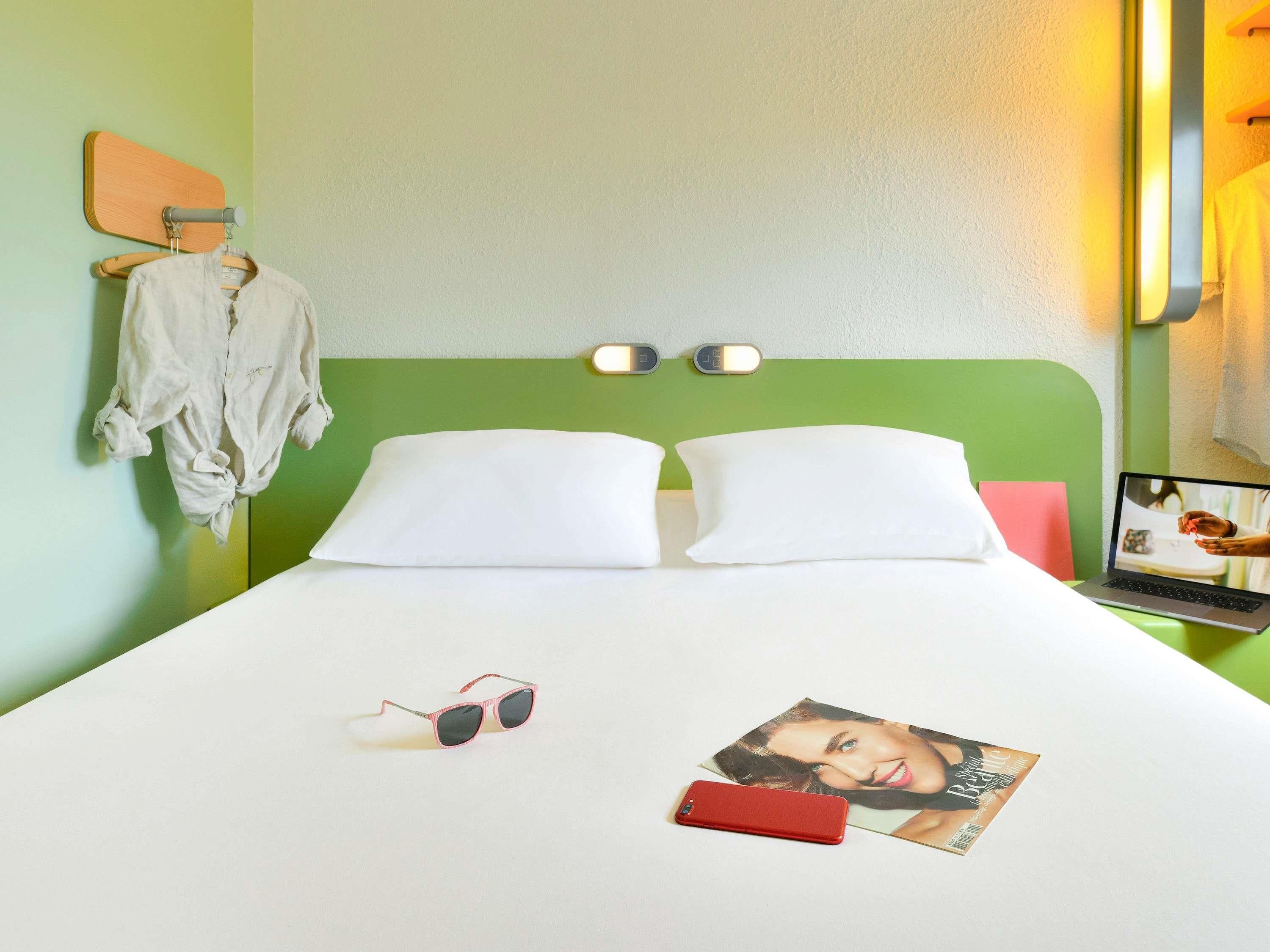 Una habitación de hotel con cabecera verde, dos almohadas blancas, una cama con sábana blanca, un par de gafas de sol, un teléfono inteligente rojo y una revista sobre la mesita de noche.