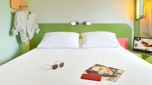 Een hotelkamer met een groen hoofdeinde, twee witte kussens, een bed met een wit laken, een paar zonnebrillen, een rode smartphone en een tijdschrift op de nachtkastje.