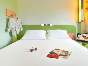 Une chambre d’hôtel avec une tête de lit verte, deux oreillers blancs, un lit recouvert d’un drap blanc, une paire de lunettes de soleil, un téléphone intelligent rouge et un magazine sur la table de chevet.