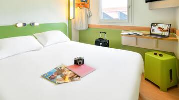 Quarto de hotel com uma cama, uma mesa de escritório, um laptop, uma mala e uma revista.