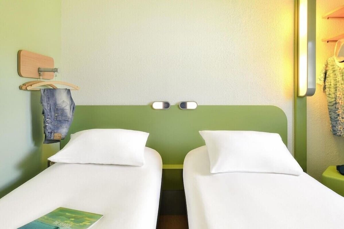 A hotel room with two single beds, a wooden headboard, a hanging garment rack, and a bedside table with a lamp.