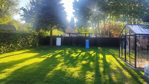 Un patio trasero bien cuidado con césped, un tobogán azul y una estructura de invernadero de vidrio.