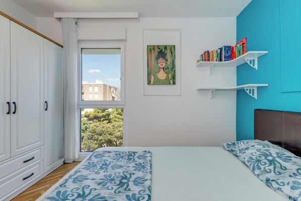 A bedroom with a bed, a window with a view, a bookshelf, and a painting on the wall.