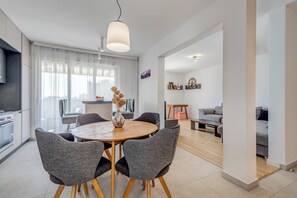 Een moderne woonkamer met een ronde houten tafel, grijze gestoffeerde stoelen, een bank en een eethoek met een witte tafel en stoelen.
