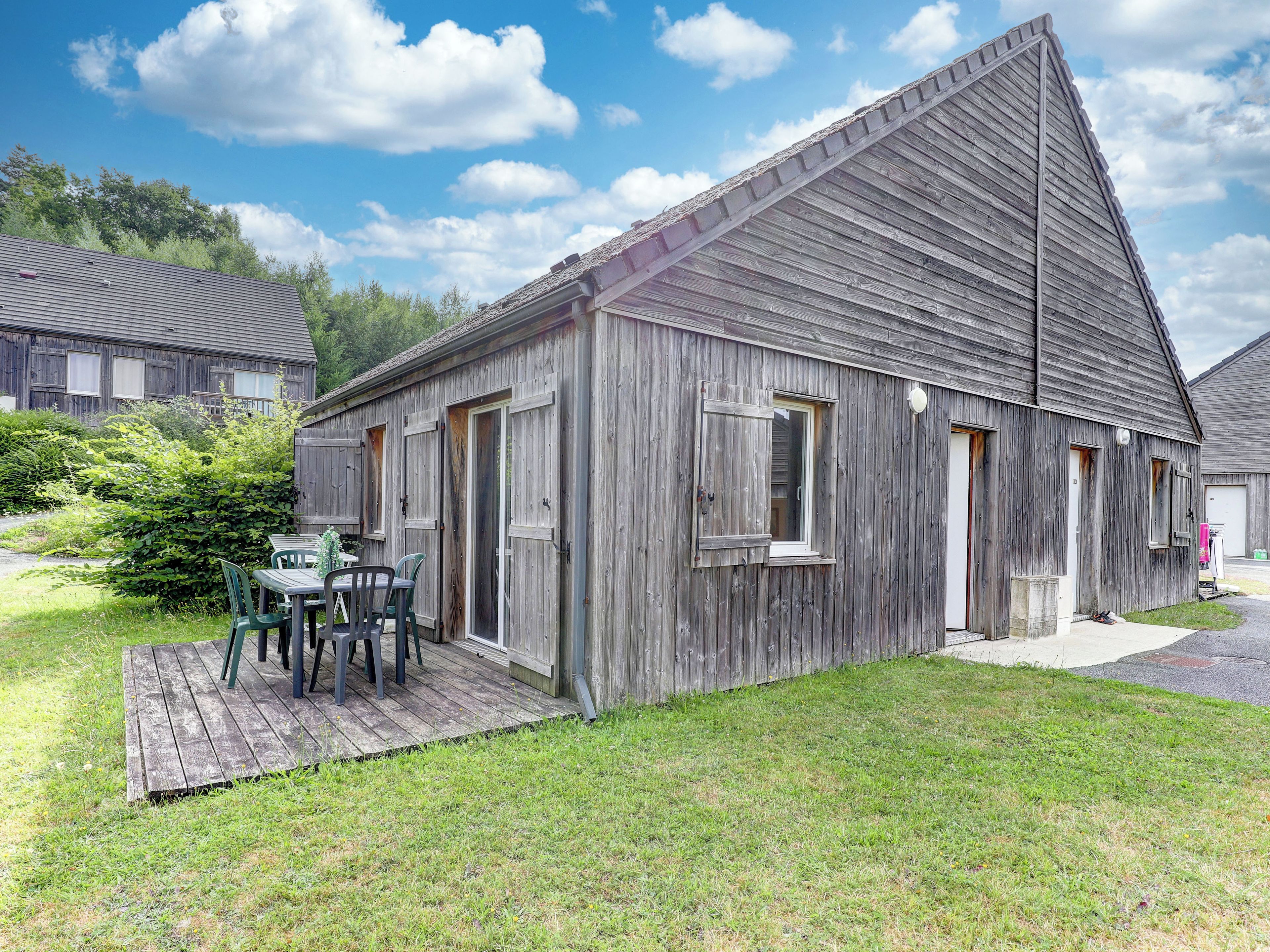 A rustic wooden building with a sloped roof, a small porch with a table and chairs, and a grassy yard.