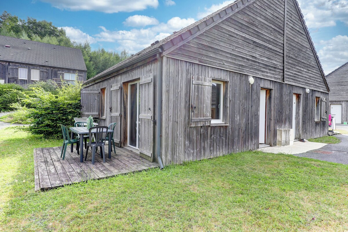 A rustic wooden building with a sloped roof, a small porch with a table and chairs, and a grassy yard.