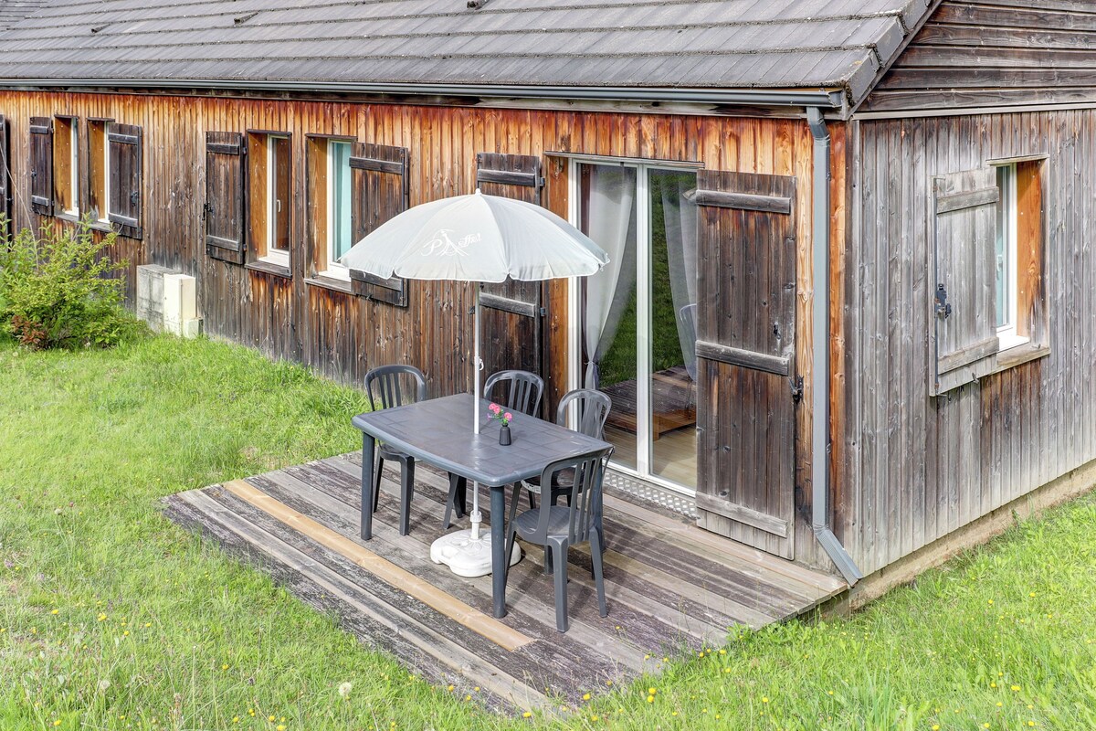 A rustic wooden building with a patio area featuring a table and chairs, a white umbrella, and a sliding glass door.