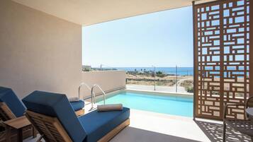 A poolside area with a view of the sea, a wooden chair, a blue lounge chair, and a pool with a clear blue sky in the background.
