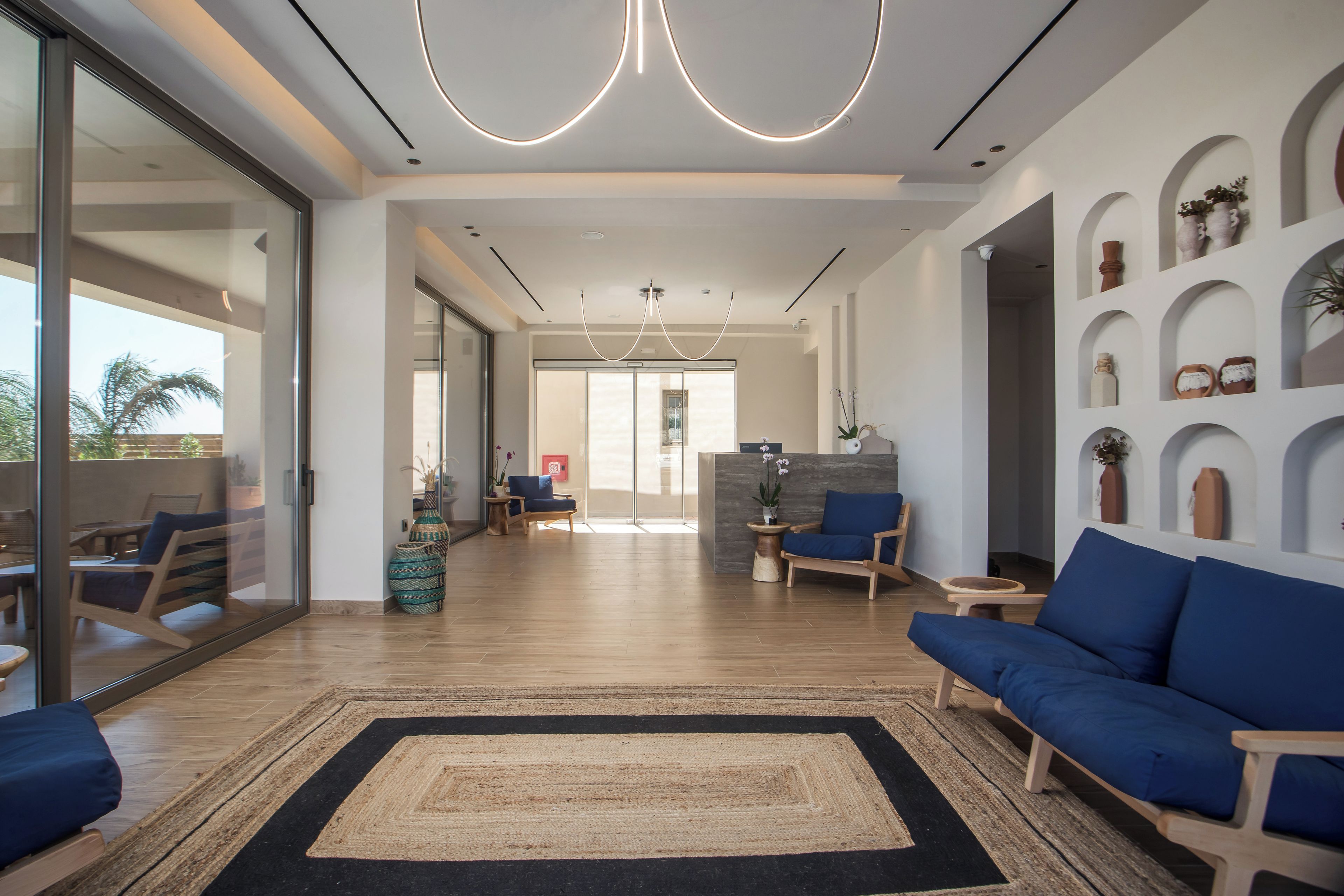 A spacious living area with a blue sofa, a coffee table, and a rug. There are potted plants and a wall with shelves displaying decorative items.