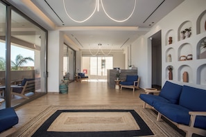 Un salón amplio con un sofá azul, una mesa de centro y una alfombra. Hay plantas en macetas y una pared con estantes que muestran objetos decorativos.