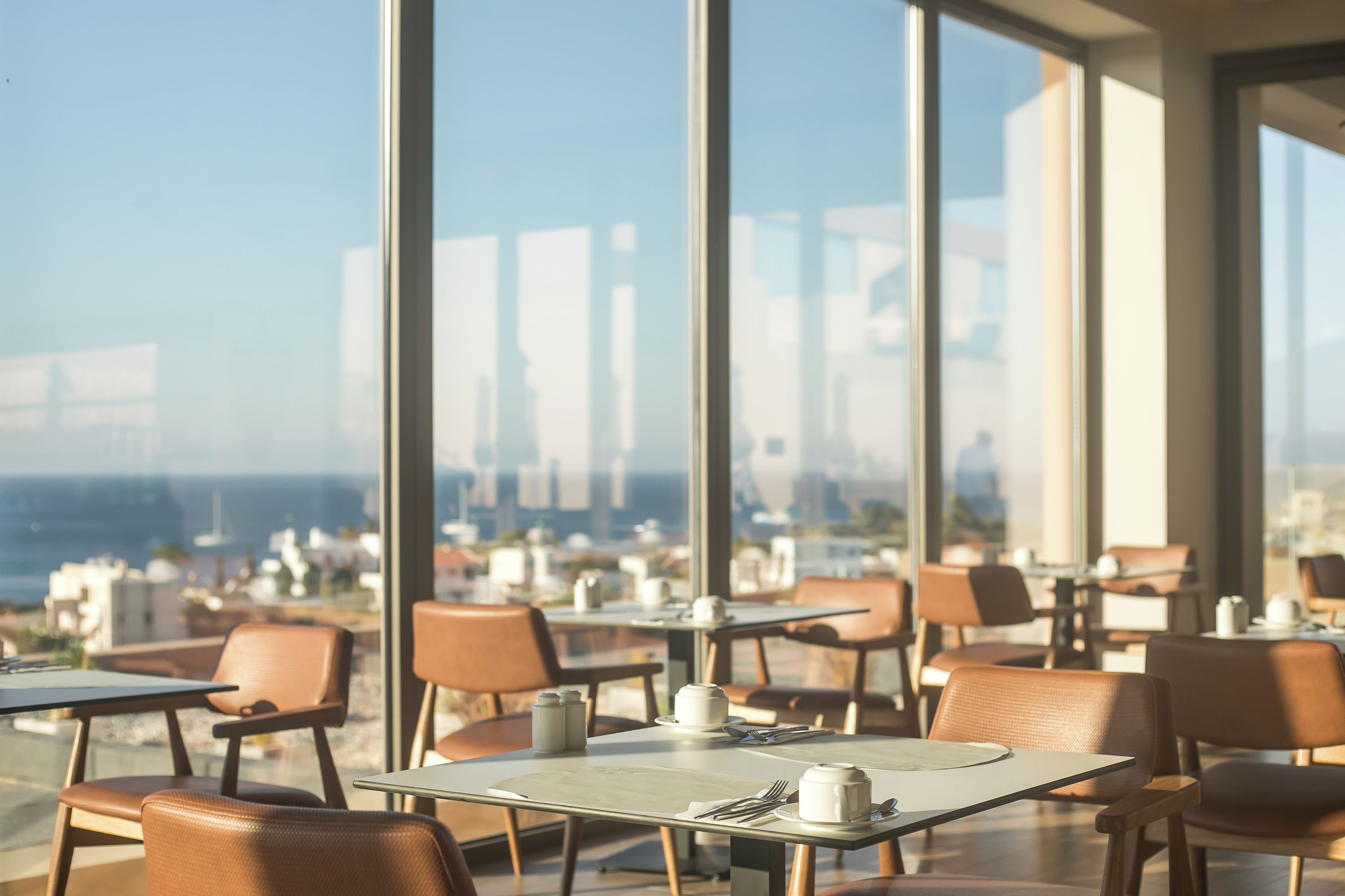 A restaurant with tables and chairs, large windows, and a view of the city and ocean.