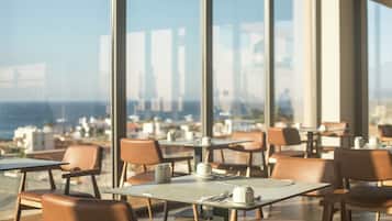 Un restaurante con mesas y sillas, amplias ventanas y vistas a la ciudad y al océano.