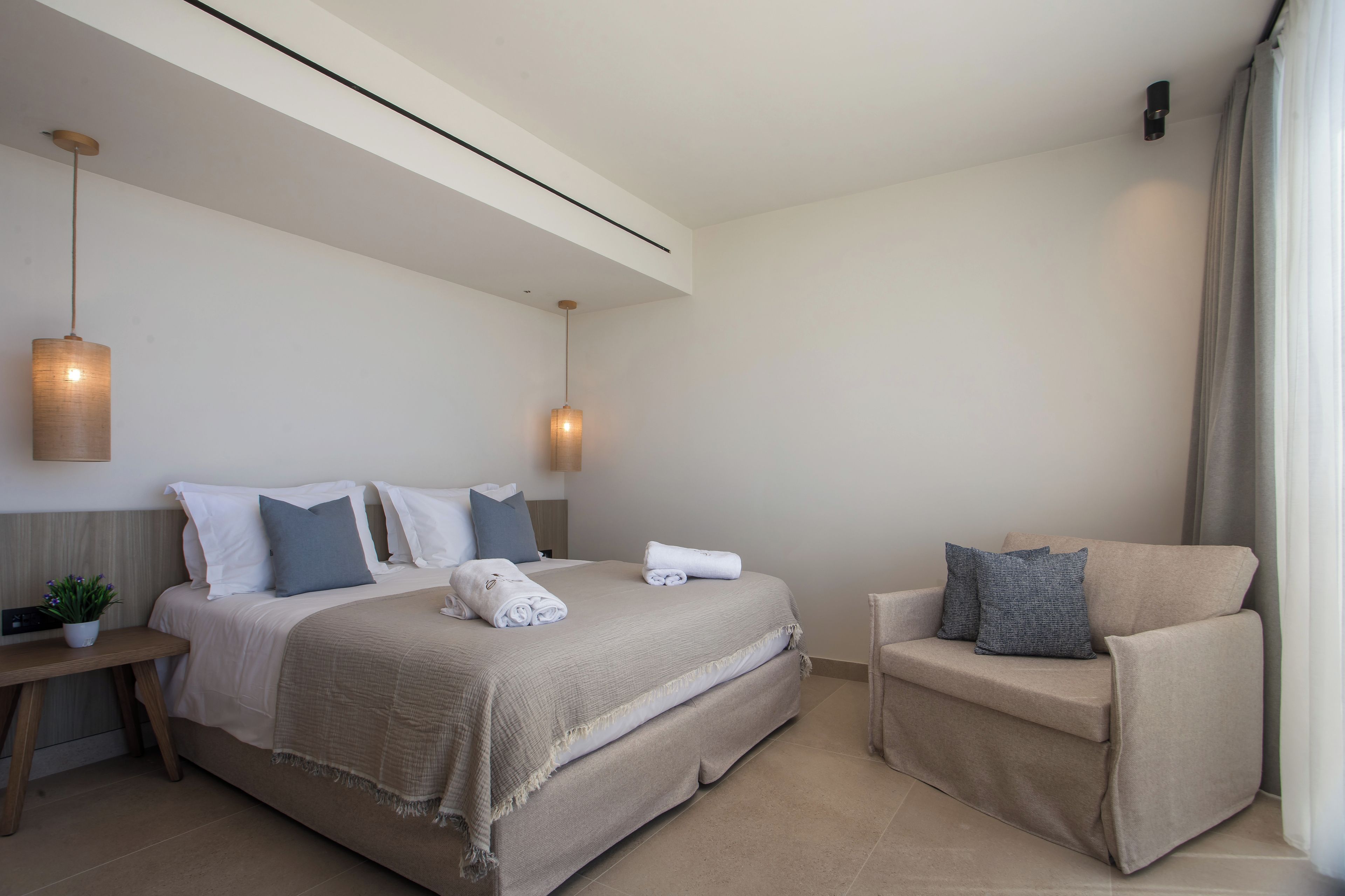 A modern hotel room with a large bed, a sofa, a bedside table with a plant, and two pendant lights.