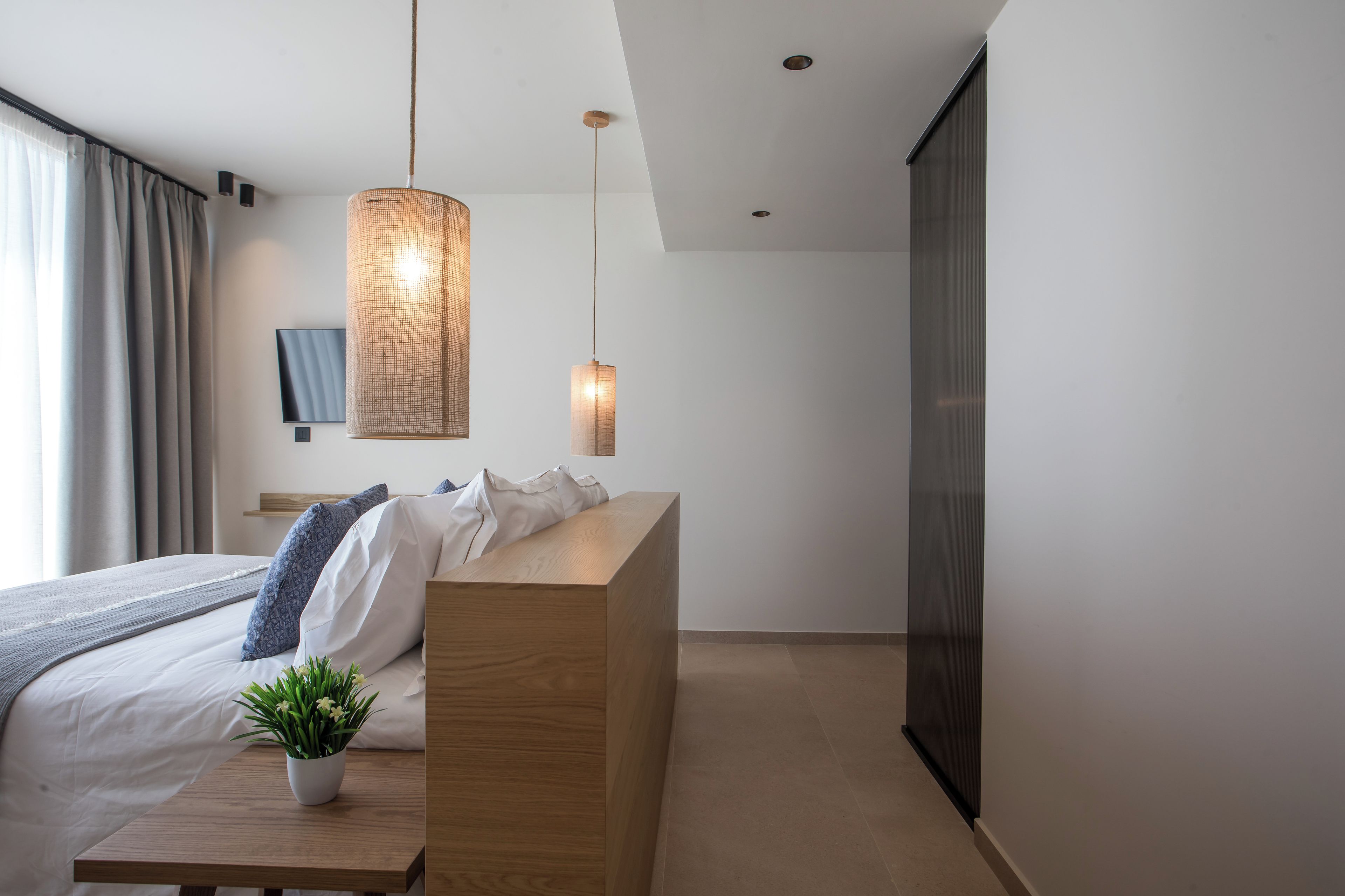 A modern bedroom with a wooden bedside table, a potted plant, a bed with white linens and blue pillows, a large woven pendant light, and a flat-screen TV mounted on the wall.