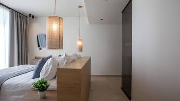 A modern bedroom with a wooden bedside table, a potted plant, a bed with white linens and blue pillows, a large woven pendant light, and a flat-screen TV mounted on the wall.