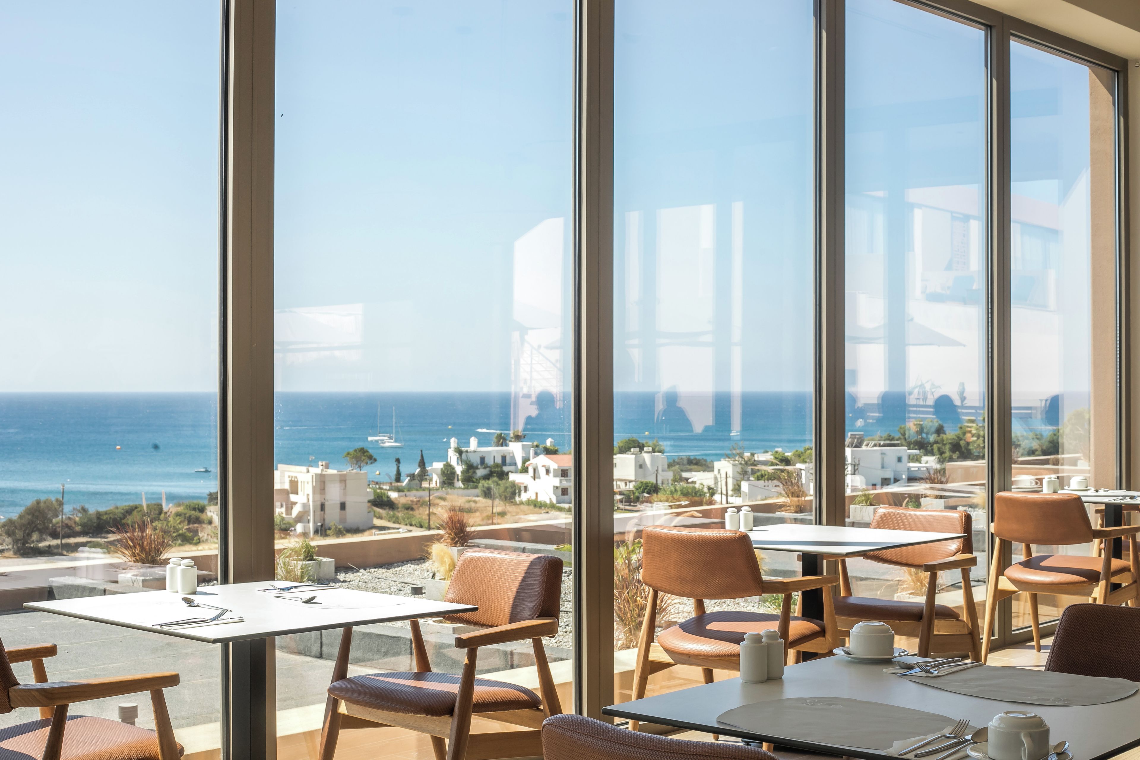A restaurant with a view of the sea and white buildings. Tables and chairs are arranged for dining.