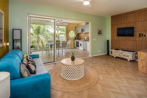A living room with a blue sofa, a round coffee table with a plant, and a kitchen area visible through a glass door.