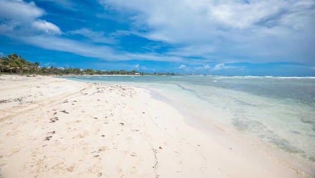 A beach with clear turquoise water, white sand, and a distant shoreline with buildings.