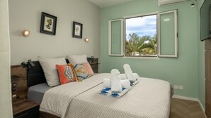 A neatly made bed with a patterned bedspread, pillows, and towels. There are two framed pictures on the wall, a window with a view of palm trees, and a bedside table with a remote control.