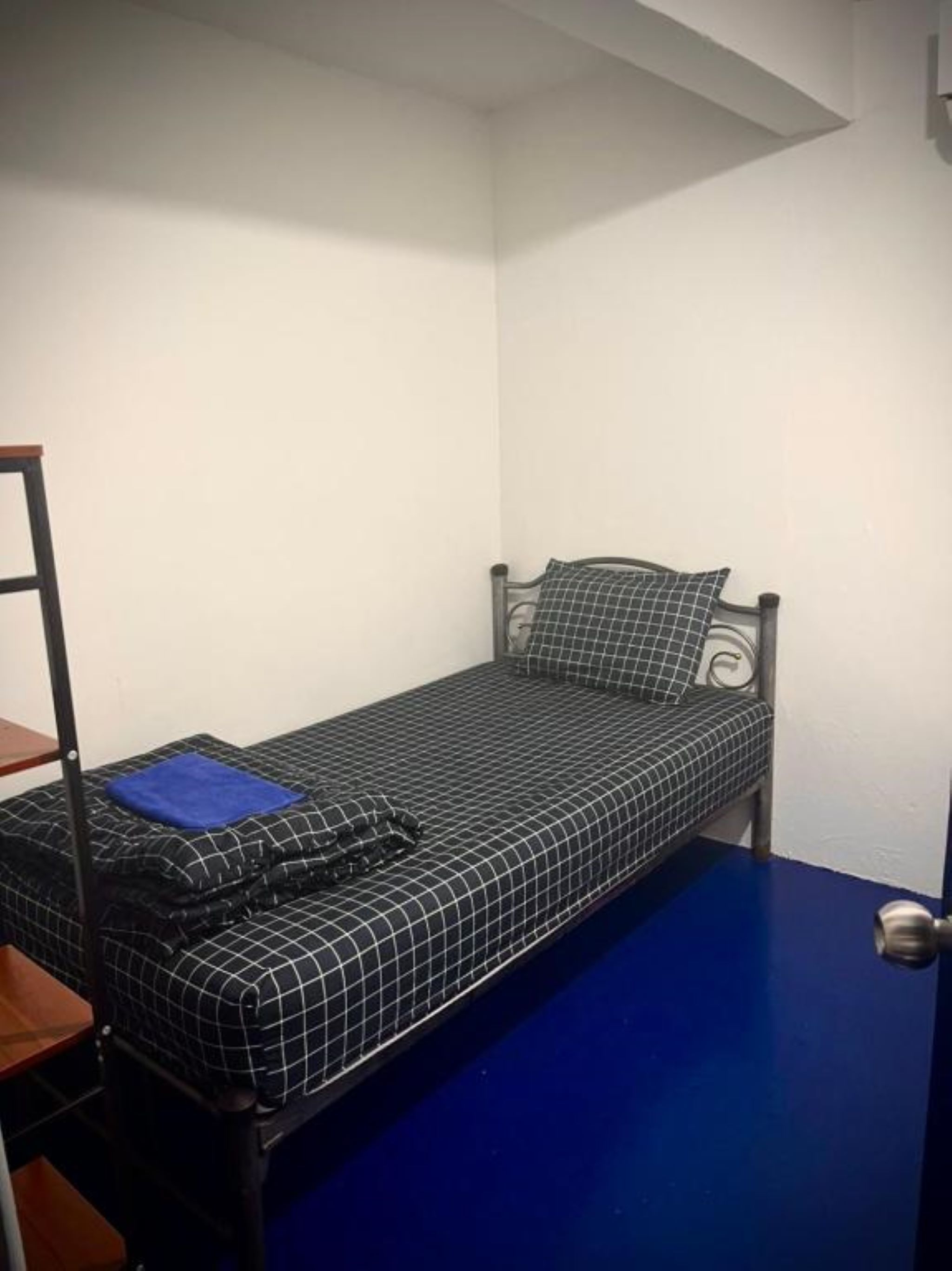 Ein kleines Zimmer mit einem Einzelbett, Metallrahmen, blauem Boden und weißer Wand.
