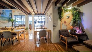 A modern cafe with wooden tables and chairs, large windows, and a mural of a face on the wall. - Arawana Hoft Hostel (Bangkok)