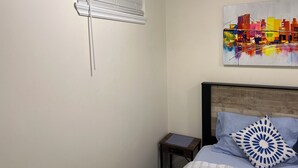 A bedroom with a bed, a nightstand, a window with blinds, and a colorful abstract painting on the wall. - Charming 1-bedroom/ kitchen n a house in beautiful Toronto with WiFi, AC, Female (Toronto)