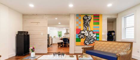 Una sala de estar con un sofá, una mesa de centro con piezas de ajedrez, un cuadro en la pared y una cocina al fondo.