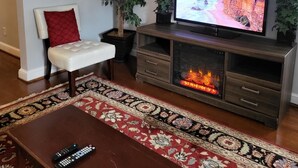 A living room with a flat-screen TV, a fireplace, a sofa, a clock, and two potted plants.