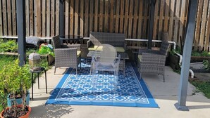 A covered patio area with outdoor furniture, a patterned rug, and a wooden fence in the background.