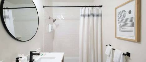 Une salle de bain moderne avec un miroir rond, un robinet noir, un lavabo blanc, un rideau de douche et une œuvre d’art abstraite encadrée accrochée au mur.