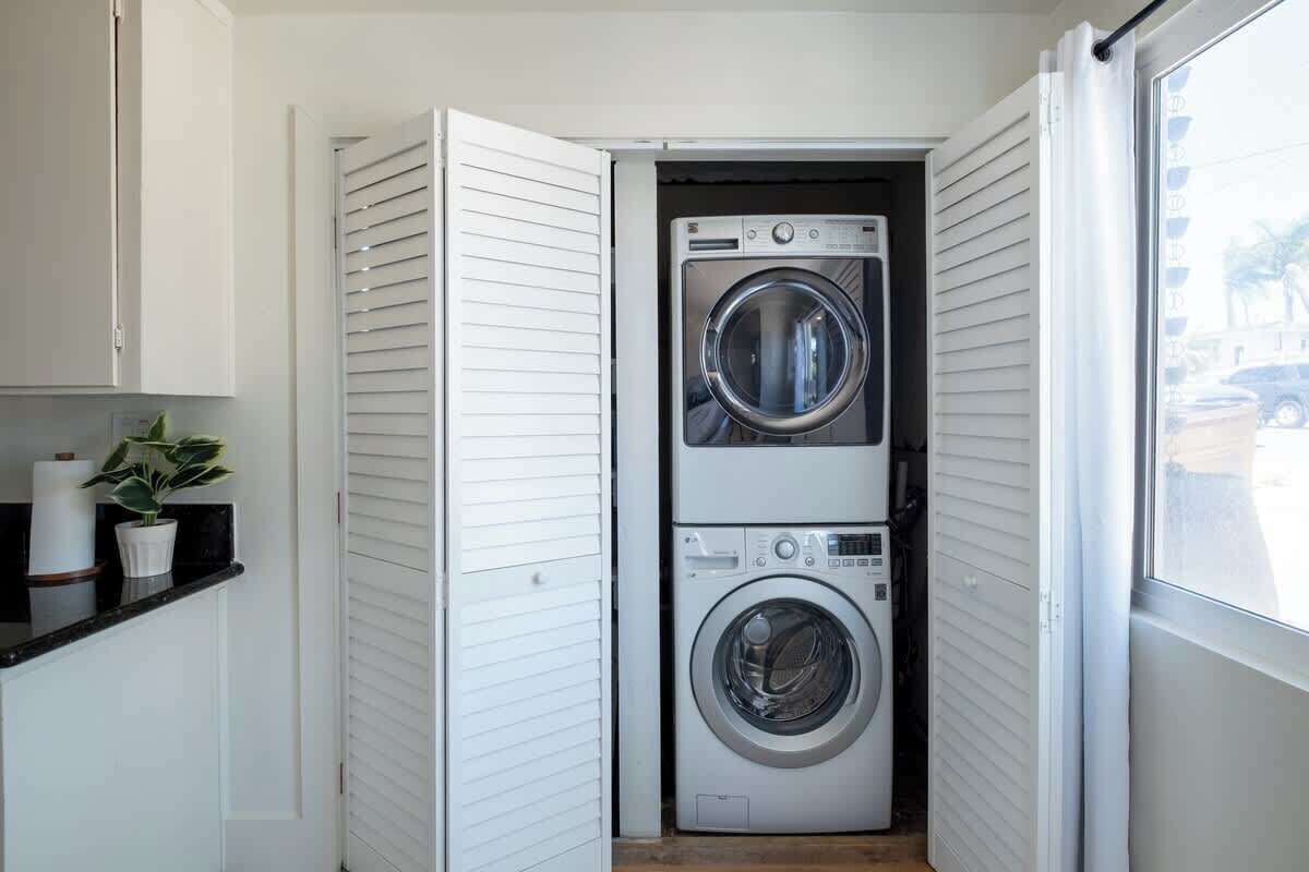 A compact laundry room with two stacked washers and dryers, a small countertop with a potted plant, and a window providing natural light.