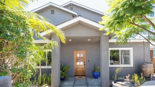 A modern house with a wooden front door, a covered porch, and a well-maintained garden with various plants and a large barrel. - Spacious Central & Chic 5BR w/ Luxe Outdoor Living (San Diego)