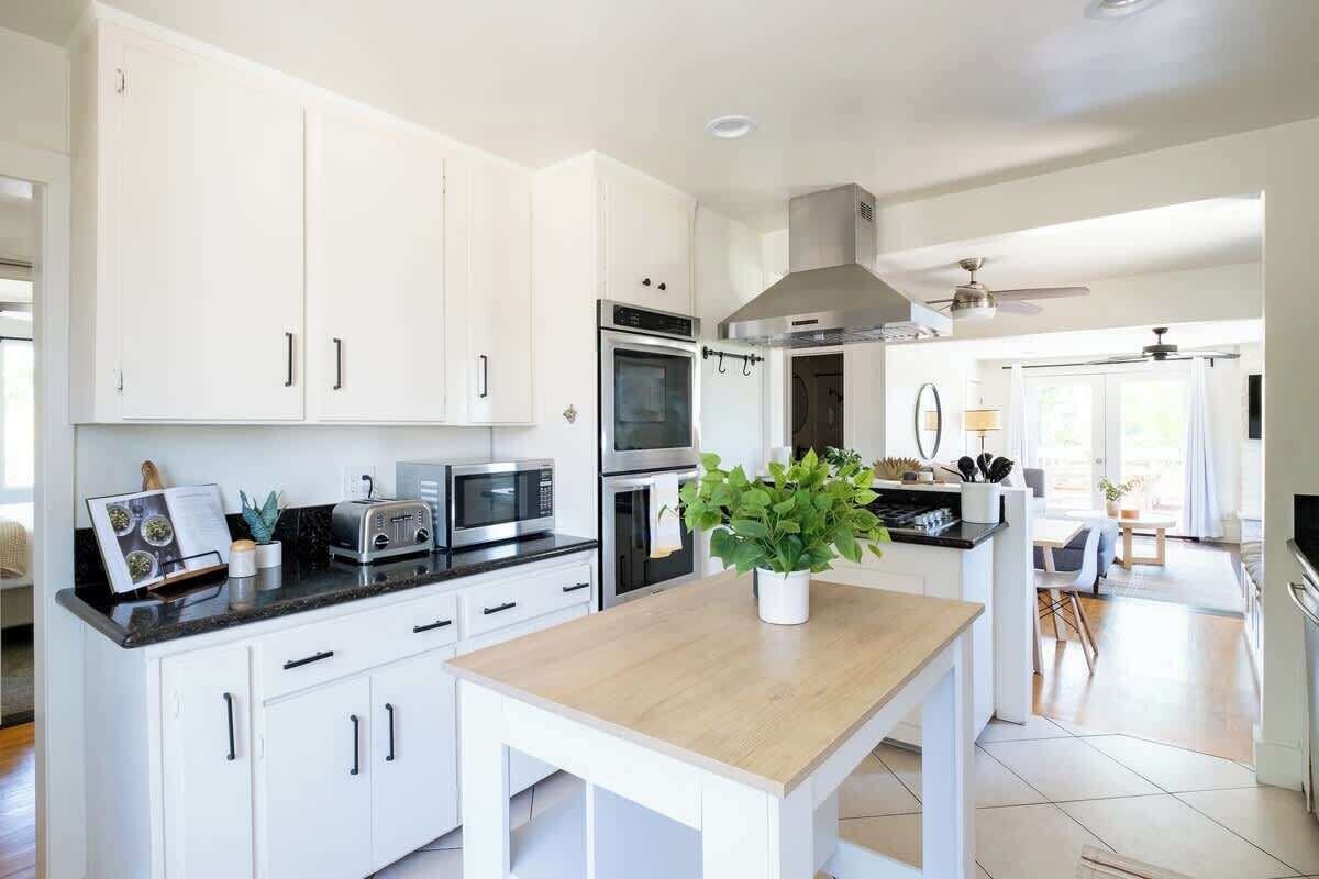 Une cuisine moderne avec des armoires blanches, un comptoir noir et une table en bois. Elle est équipée d’appareils électroménagers intégrés, notamment un four à micro-ondes, un four et une cuisinière. Une plante en pot est posée sur la table, et la cuisine donne sur un coin repas avec vue sur l’extérieur.