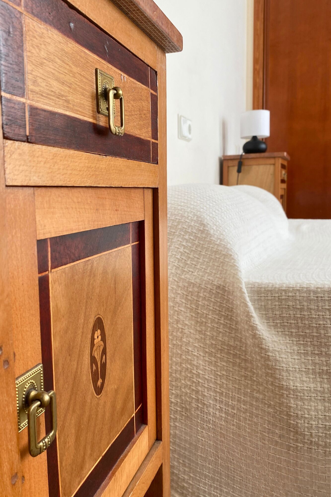 Ein Holzschrank mit Messingschloss und -griff, ein Bett mit beiger Decke und ein Nachttisch aus Holz mit einer Lampe.