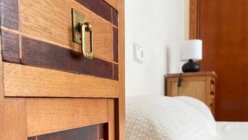 Ein Holzschrank mit Messingschloss und -griff, ein Bett mit beiger Decke und ein Nachttisch aus Holz mit einer Lampe.