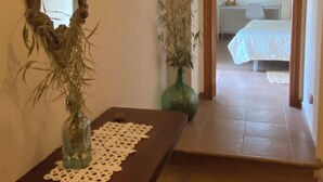 Un pasillo con una mesa de madera, un espejo y un jarrón verde con plantas secas. Se ve una cama en la habitación contigua.