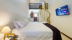 Habitació d'hotel amb un llit, una televisió penjada a la paret, una taula de nit amb una làmpada i una escala que porta a un nivell superior.