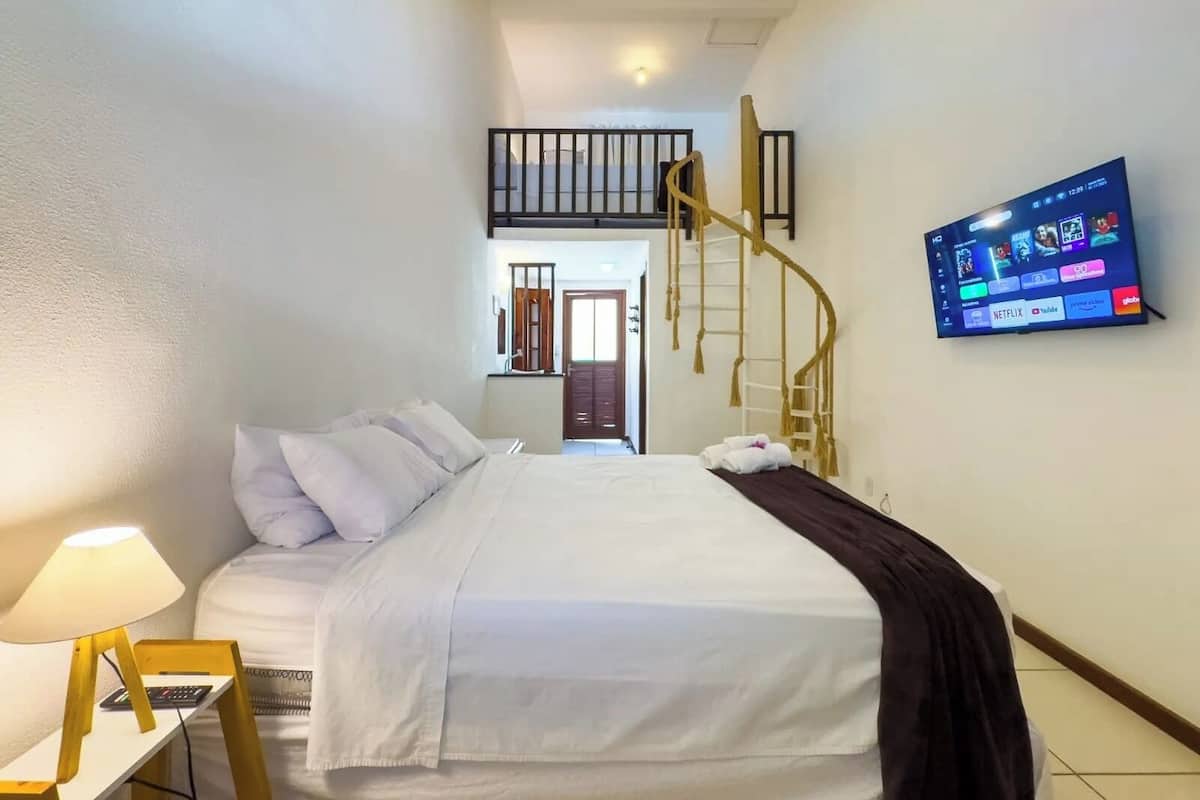 A hotel room with a bed, a TV mounted on the wall, a desk with a lamp, and a staircase leading to an upper level.
