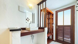 Bany amb un lavabo blanc, prestatges de fusta i una porta amb persianes horitzontals.