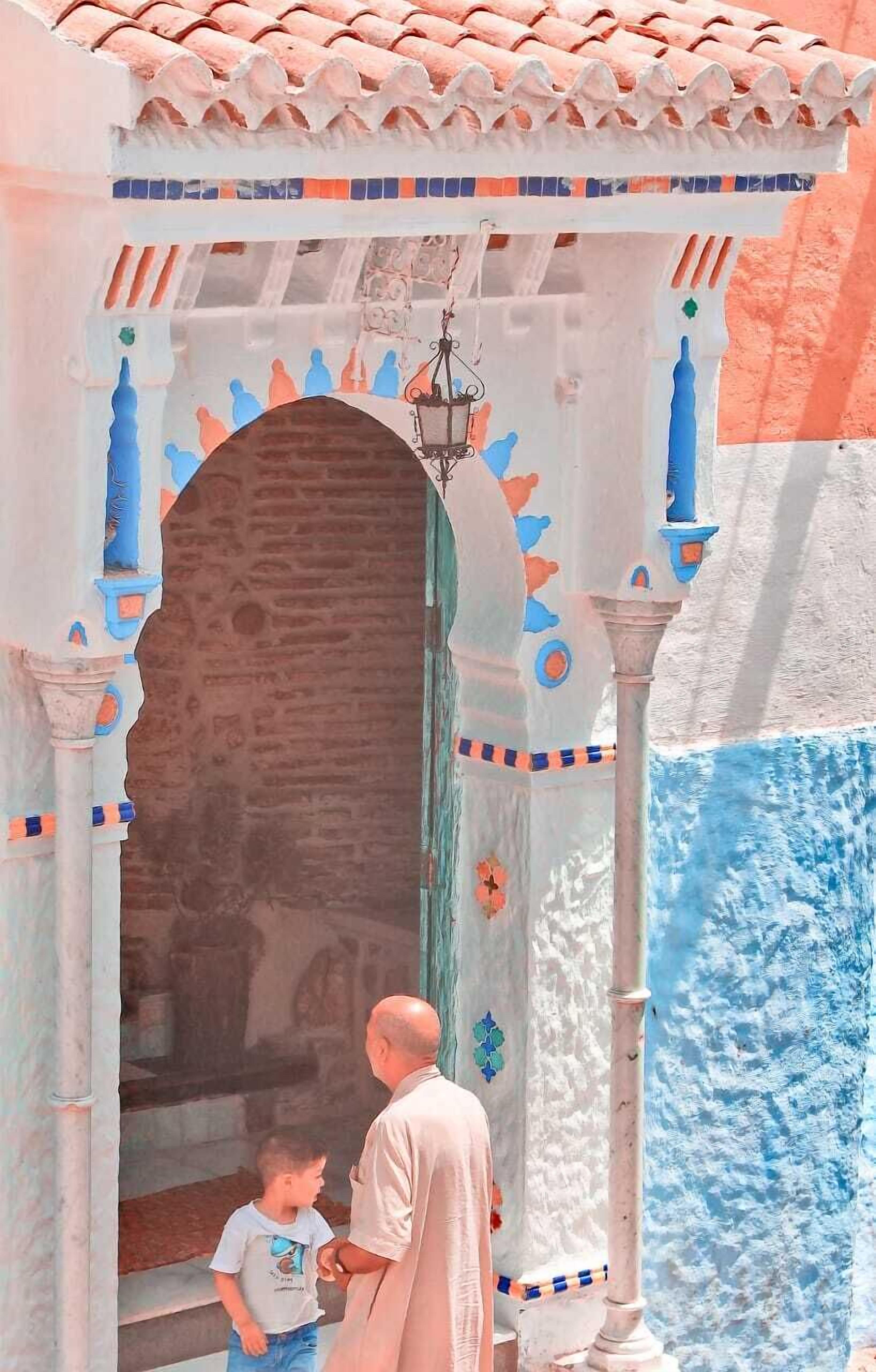 A man and a child standing at the entrance of a building with a tiled roof and a decorative archway.
