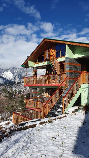 A multi-story building with wooden balconies and stairs, set against a snowy mountain backdrop. - Bohodyssey Hallan (Kullu)