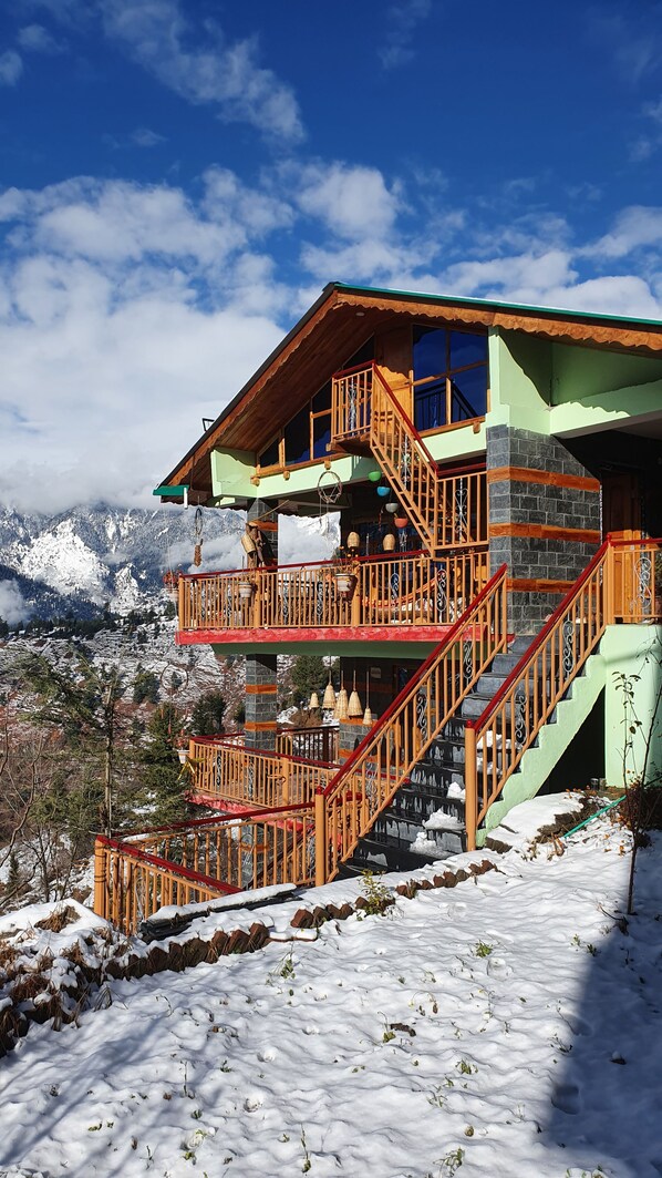 A multi-story building with wooden balconies and stairs, set against a snowy mountain backdrop. - Bohodyssey Hallan (Kullu)
