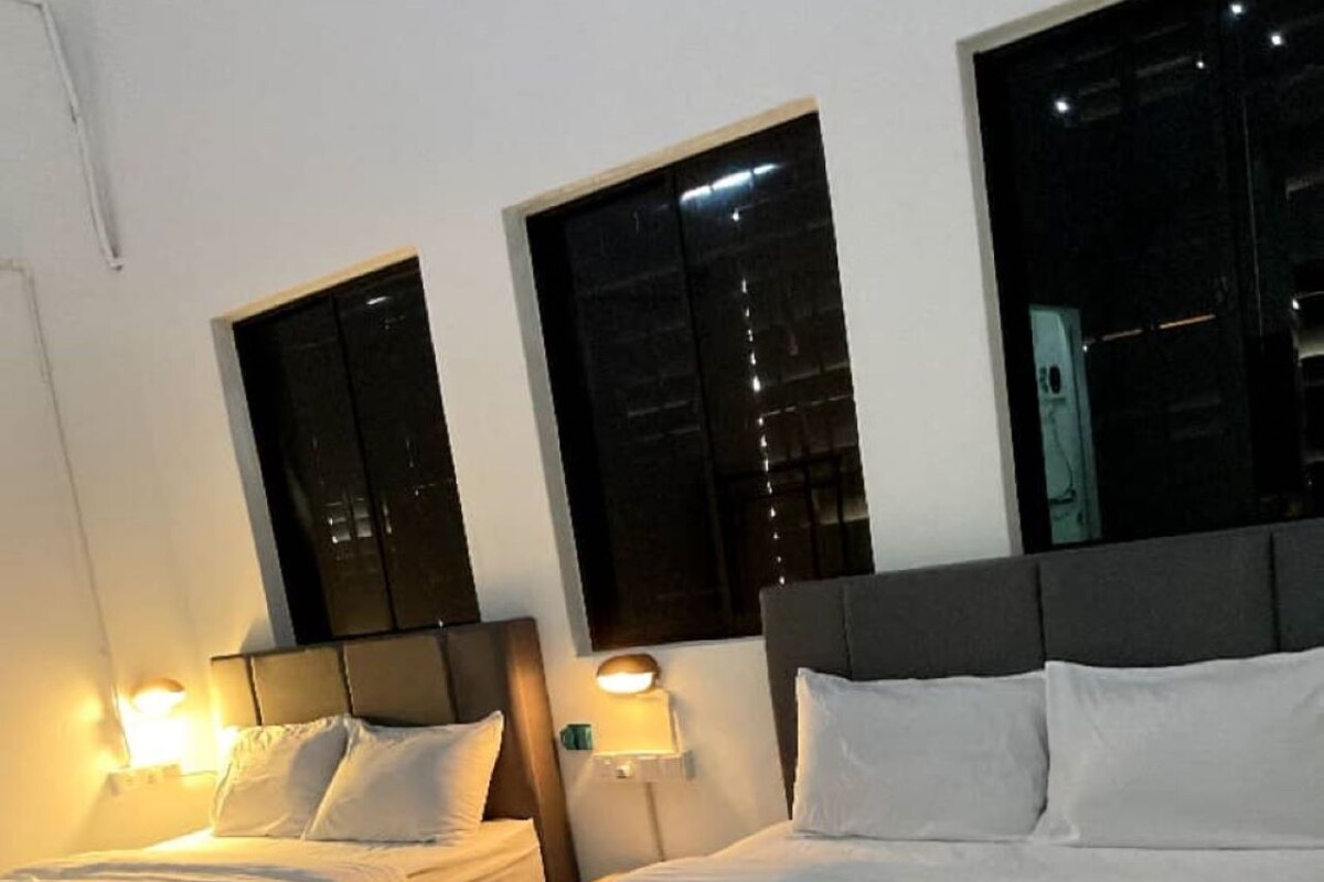 A hotel room with two single beds and one double bed, each with white linens and pillows. There are two bedside tables with lamps, and the room has a modern design with a grey headboard and a large window.