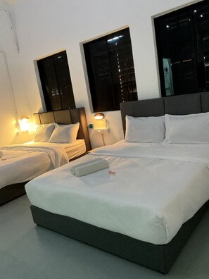 Ein Hotelzimmer mit zwei Einzelbetten und einem Doppelbett, jeweils mit weißen Bettwäsche und Kissen. Zwei Nachttische mit Lampen stehen bereit, und das Zimmer überzeugt durch ein modernes Design mit grauem Kopfteil und einem großen Fenster.
