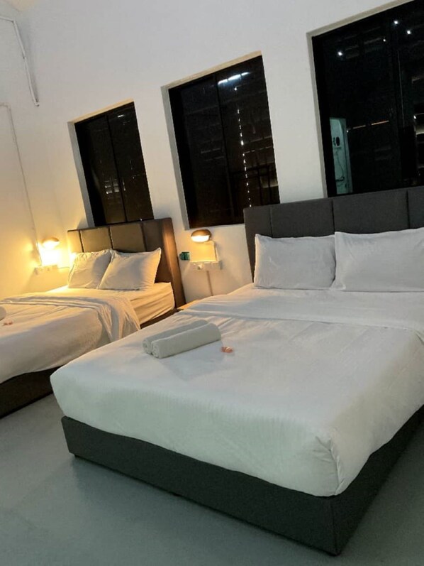Quarto de hotel com duas camas de solteiro e uma cama de casal, cada uma com roupa de cama e almofadas brancas. Possui duas mesas de cabeceira com abajures, e o quarto apresenta um design moderno com cabeceira cinza e uma grande janela.