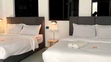 Two single beds with grey headboards and white bedding, a bedside table with a lamp, and large windows with night views.