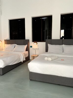 Duas camas de solteiro com cabeceiras cinzentas e roupa de cama branca, uma mesa de cabeceira com abajur, e janelas amplas com vista noturna.