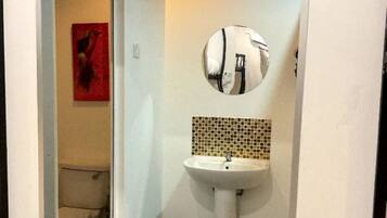A small bathroom with a white sink, a round mirror, and a toilet visible through an open door.